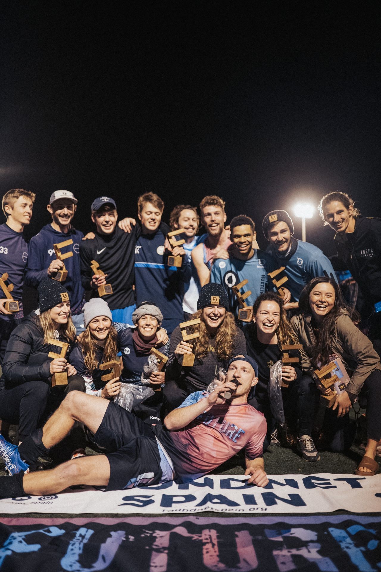 Group of people posing with trophies on a sports field at night, smiling and celebrating.
