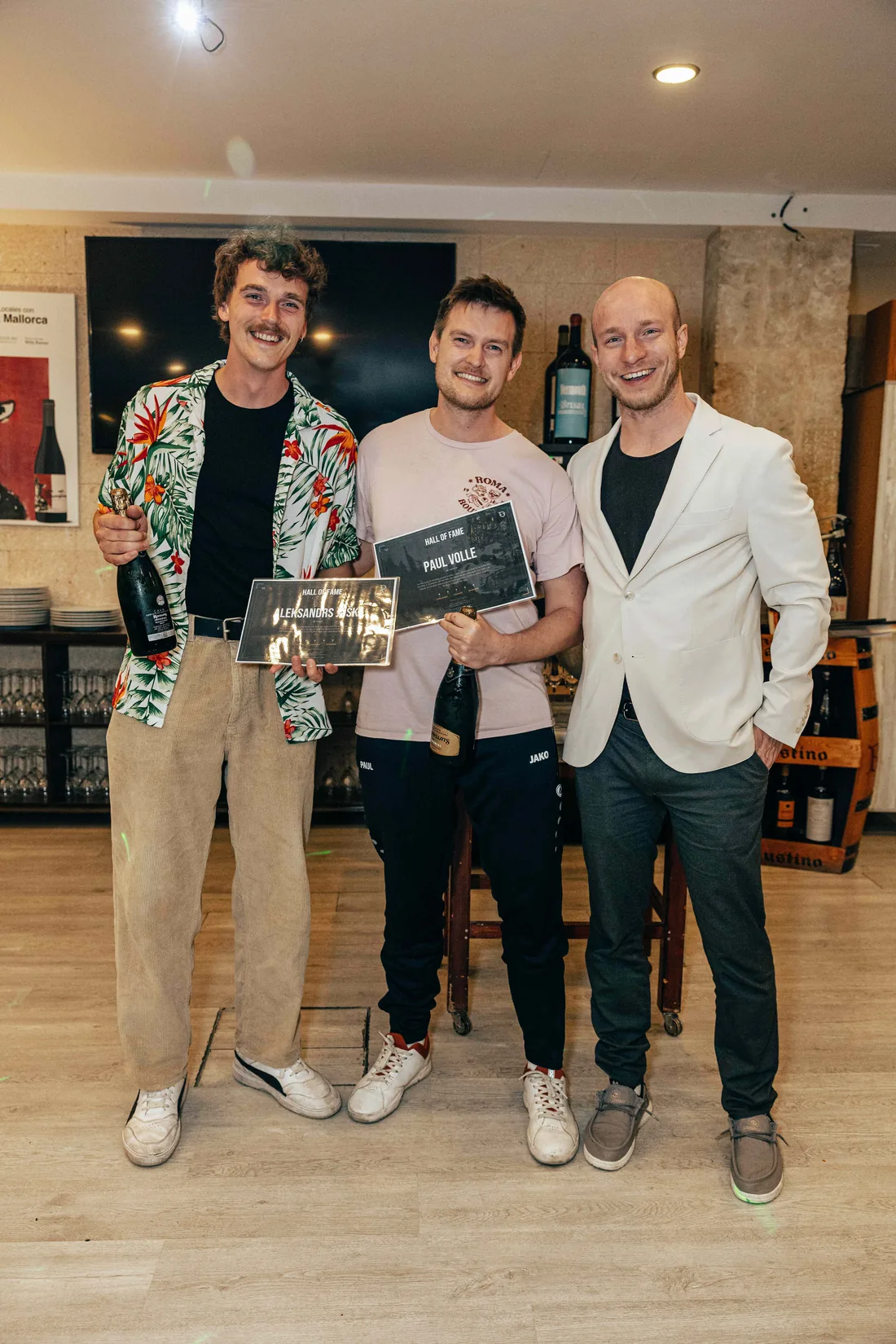 Three men holding framed Hall of Fame certificates indoors, two of them also holding champagne bottles.