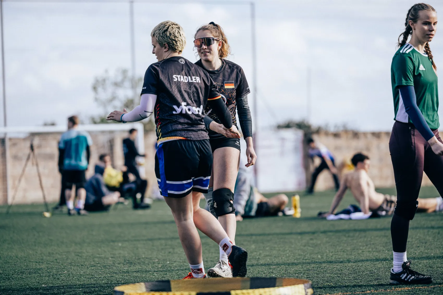 Two roundnet players in German national kit standing close together between points, one in sunglasses, other players and the field behind them.