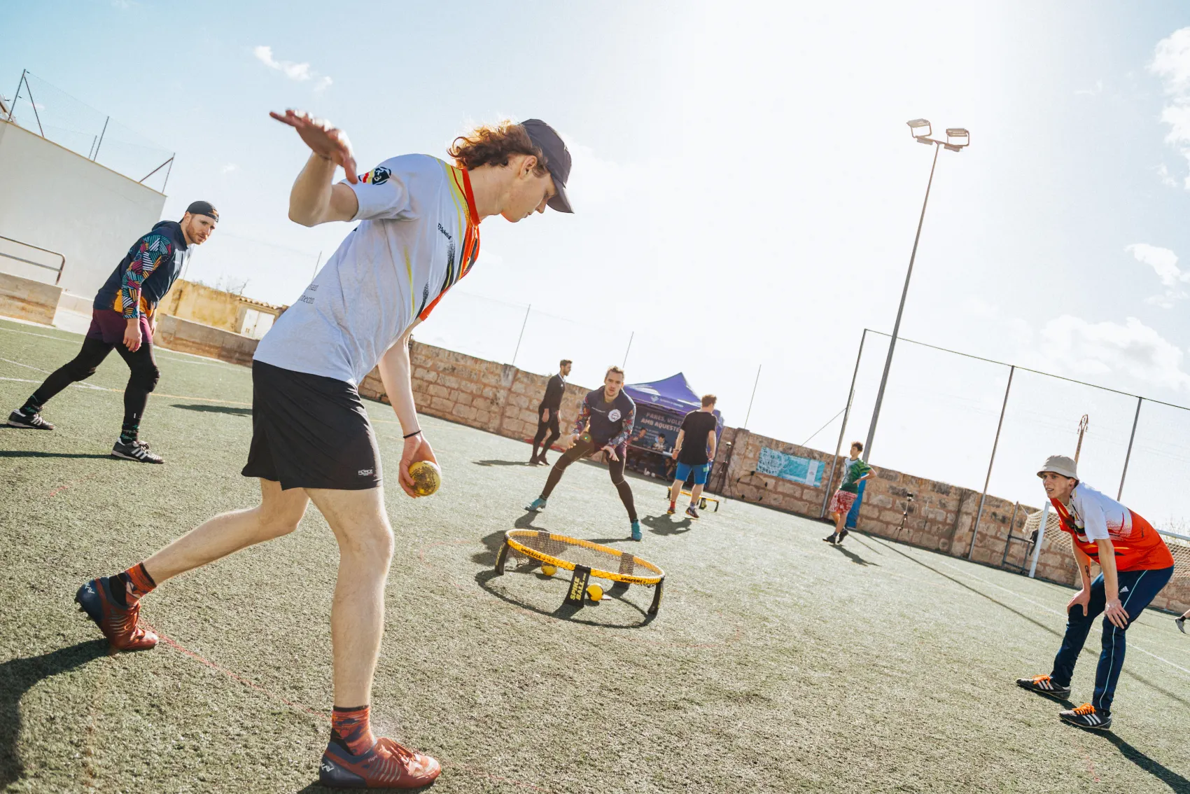 A roundnet player striding into a serve with the ball in his hand, opponents waiting around the net on an open field.