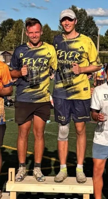 Connor Nelson and Kieran Rose in FEVER Roundnet kit on the first-place step of a podium, each holding a trophy at an outdoor tournament.