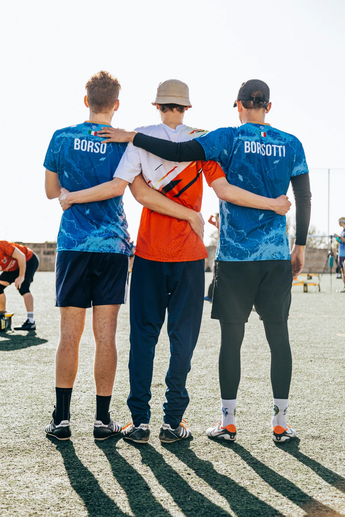 Three roundnet players standing arm in arm with their backs to the camera, two of them in blue Italian national jerseys.