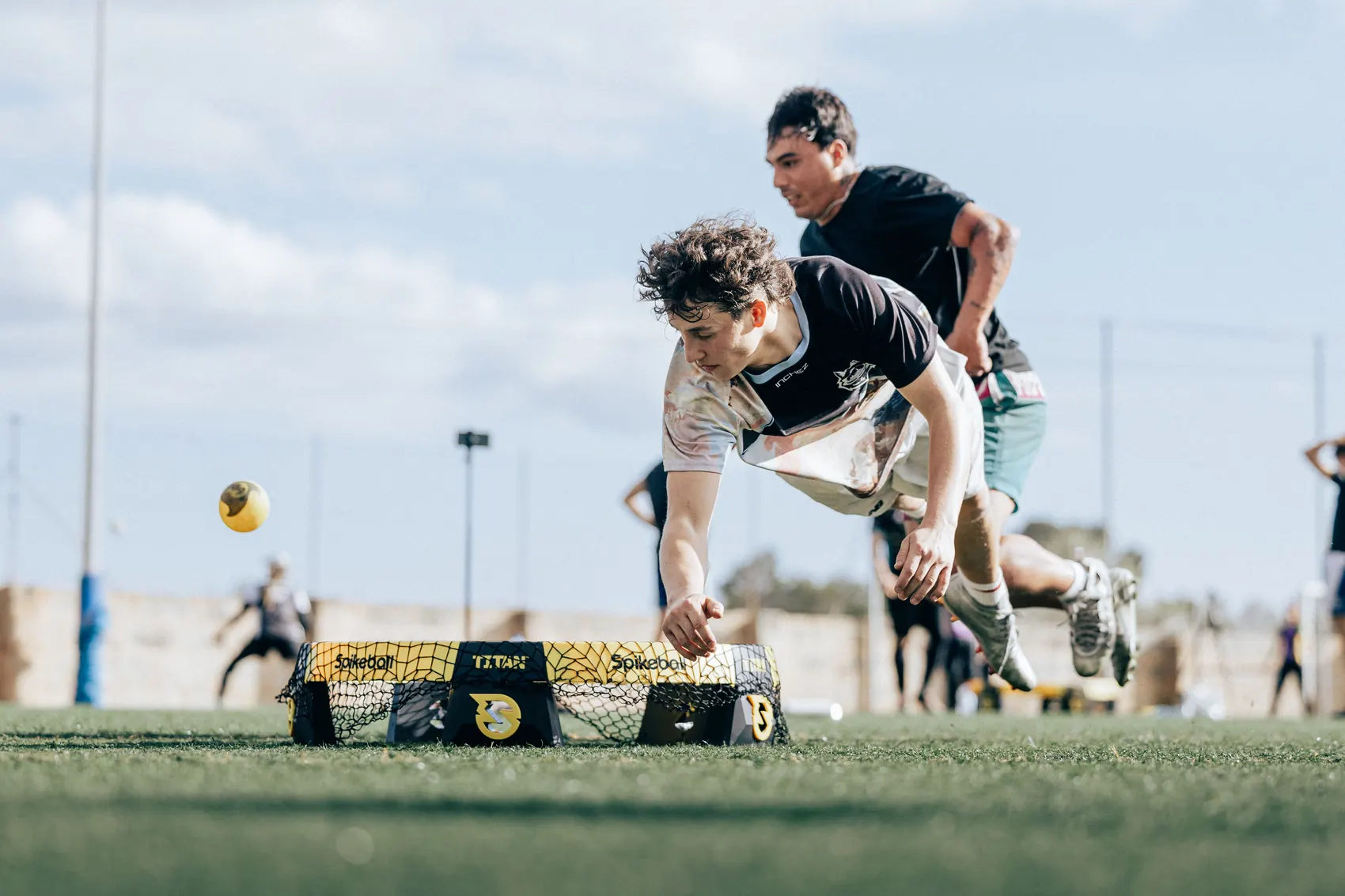 A roundnet player diving full stretch to reach a yellow ball just above the grass, an opponent sprinting in behind him.